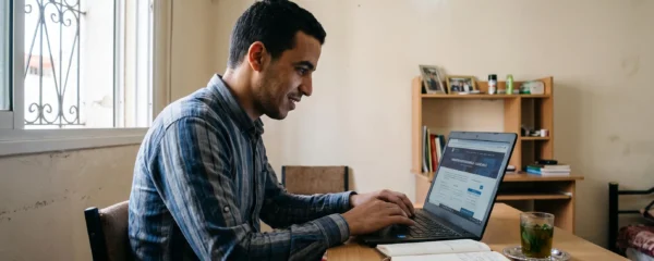 Un jeune homme marocain recherche une formation professionnelle sur un ordinateur portable, assis à son bureau à la lumière naturelle d'un appartement urbain.